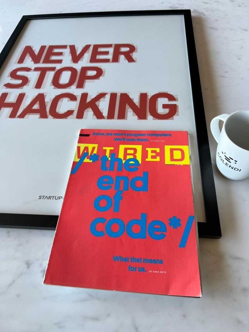 A framed 'Never Stop Hacking' print, a red WIRED magazine issue titled 'the end of code', and a white Colendi mug on a marble desk.
